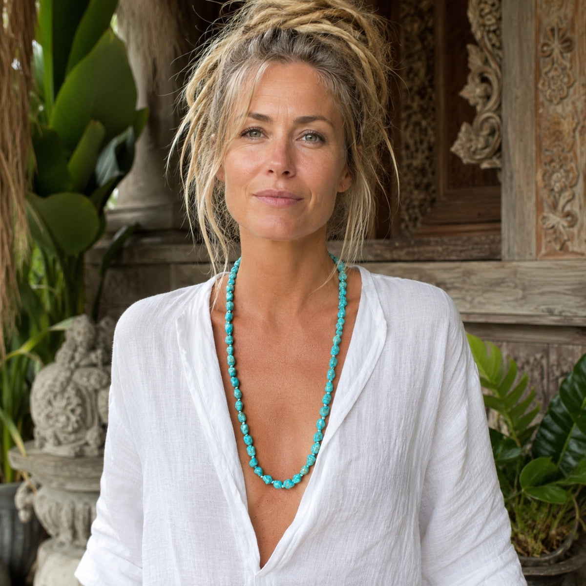 Woman wearing a handmade Bali-inspired blue stone bead necklace with a white linen blouse in a lush bohemian setting. 