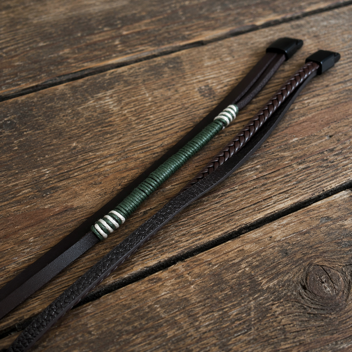 Two dark brown casual bracelets with braided texture, green wrapped cord and black metallic details on a wooden surface.