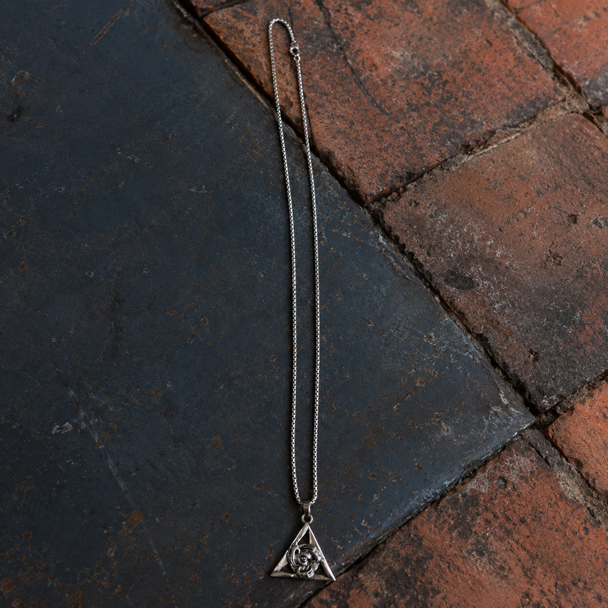 Silver-tone triangle eye pendant necklace displayed on dark metal and red brick background