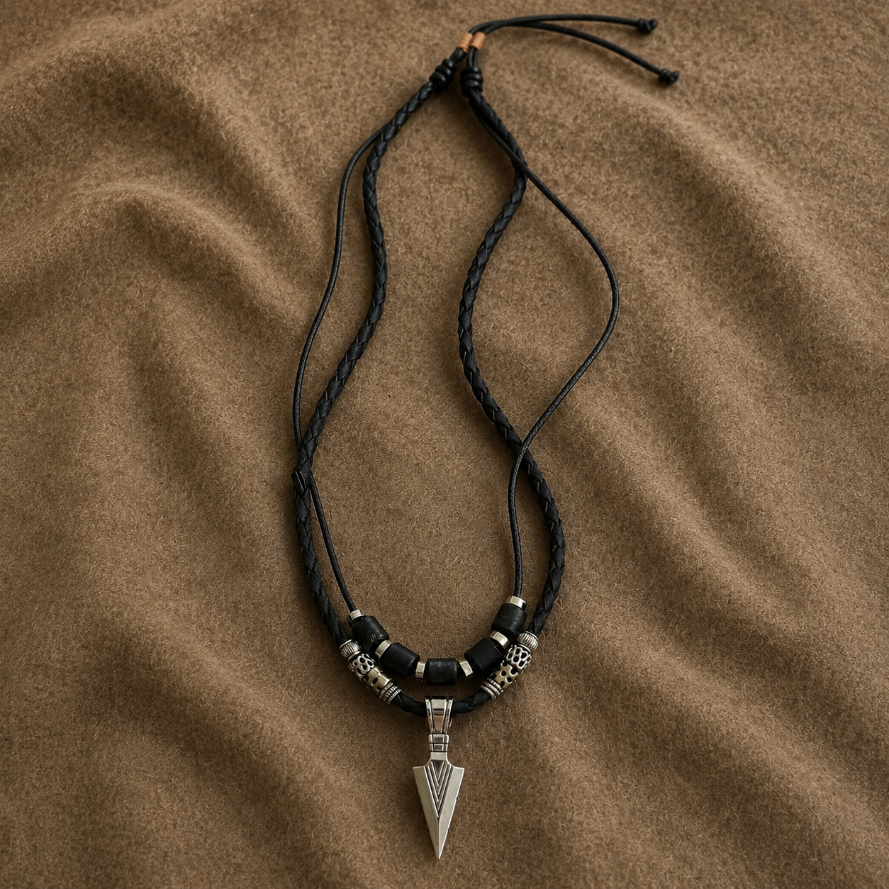 A braided black cord necklace with silver-toned metal beads and a silver arrowhead pendant, laid flat on a textured brown fabric background.