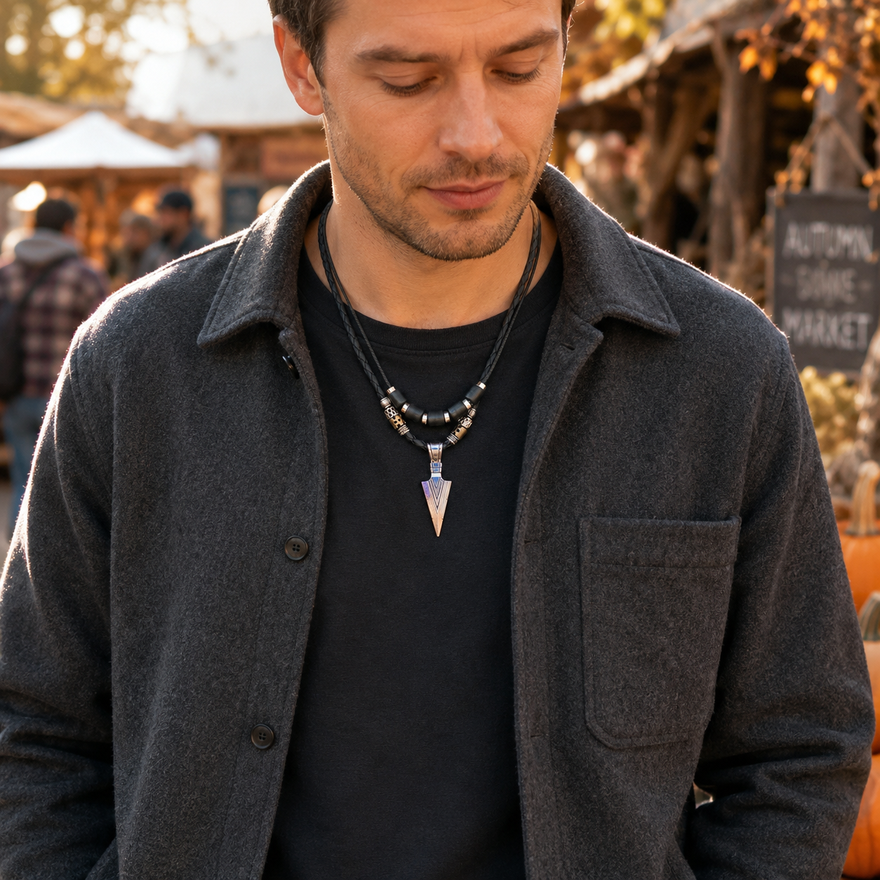 A man wearing a dark charcoal gray button-up overshirt over a black t-shirt, showcasing a layered necklace with a silver-toned beaded chain and an arrow shaped metallic pendant.