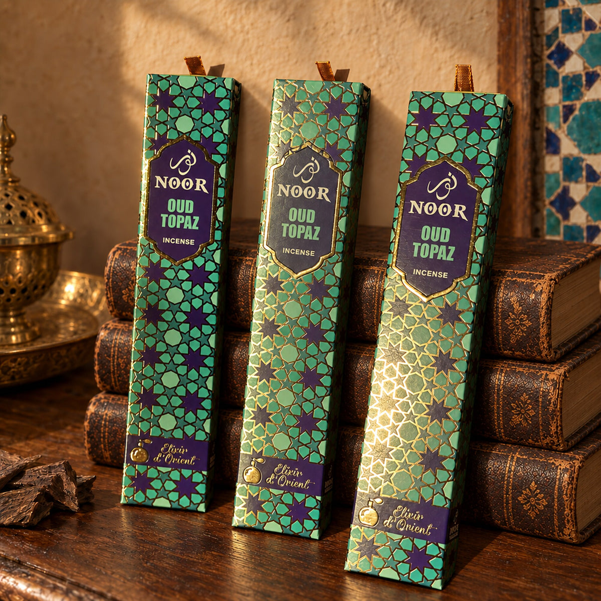 Noor Oud Topaz incense boxes in turquoise, violet and gold packaging displayed beside decorative books and brass décor.