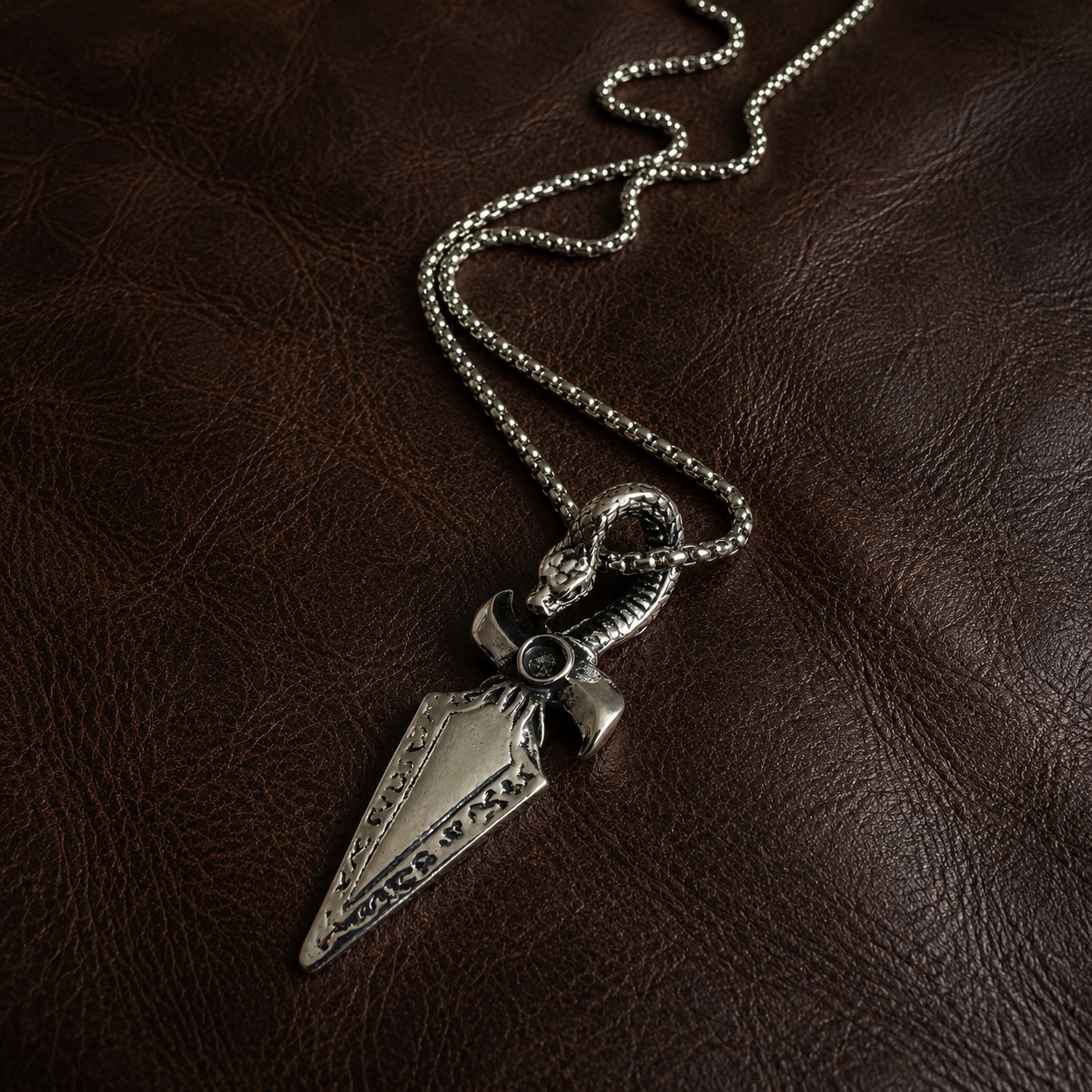 Stainless steel serpent and dagger pendant necklace with chain displayed on dark brown leather.
