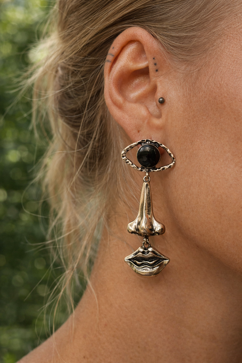 Nomad style gold earrings worn on ear. Model wearing surreal face drop earrings with an eye-shaped top, black oval cabochon, nose charm and lips charm.