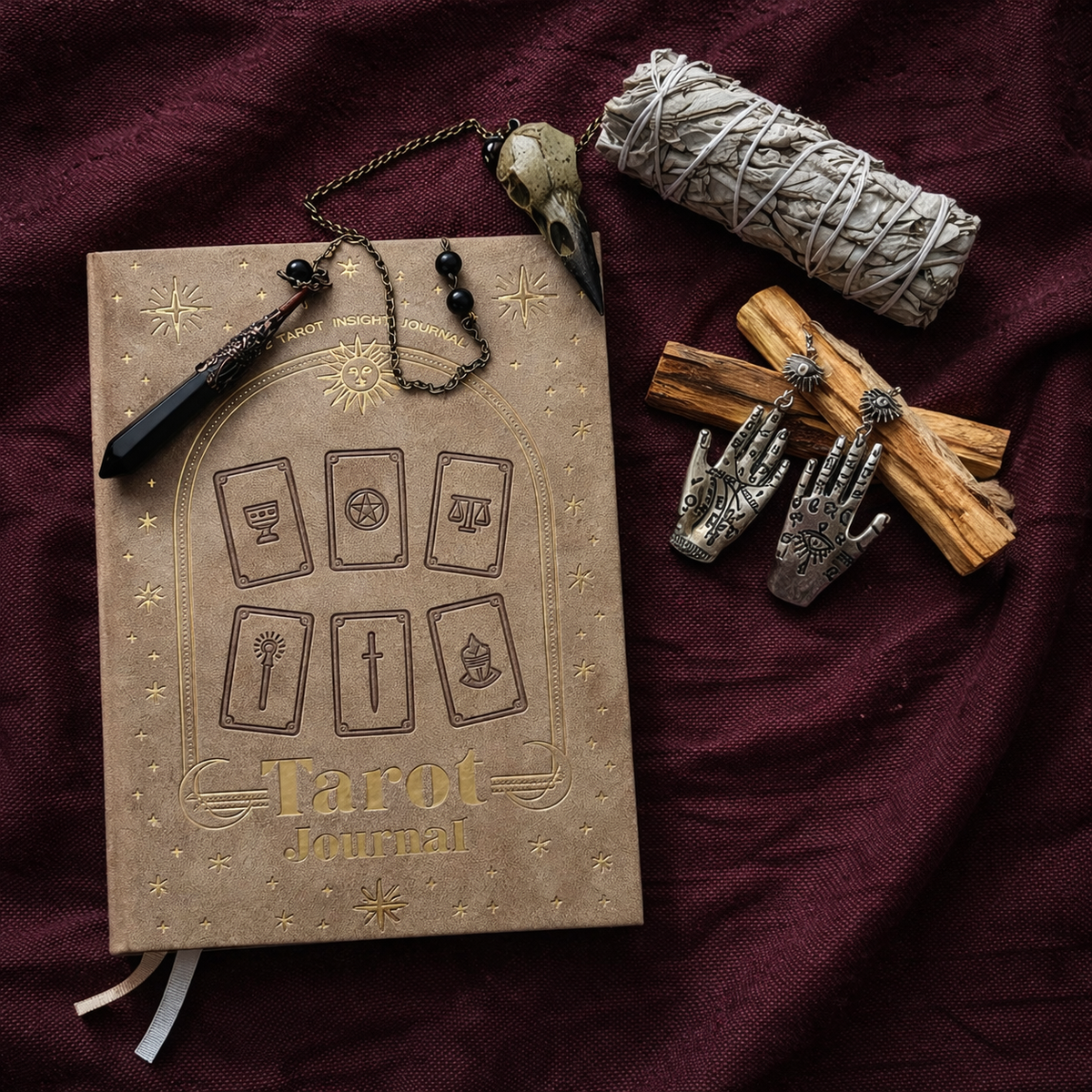 Beige Tarot Companion Journal with gold celestial motifs and tarot-card illustrations, photographed with decorative accessories on burgundy fabric